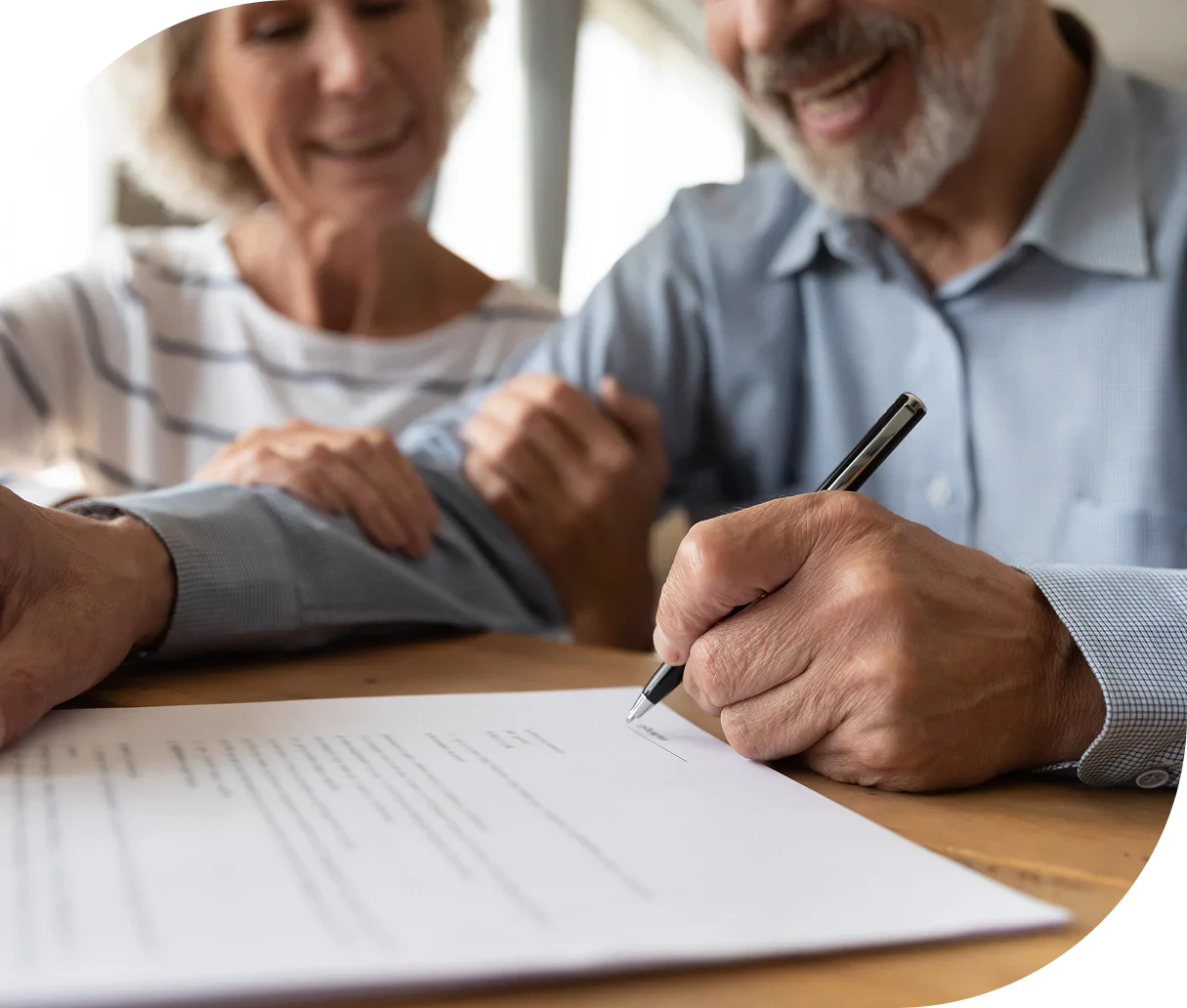 An elderly man is signing a document on a table, with a woman gently resting her hand on his arm, showcasing a moment of support.