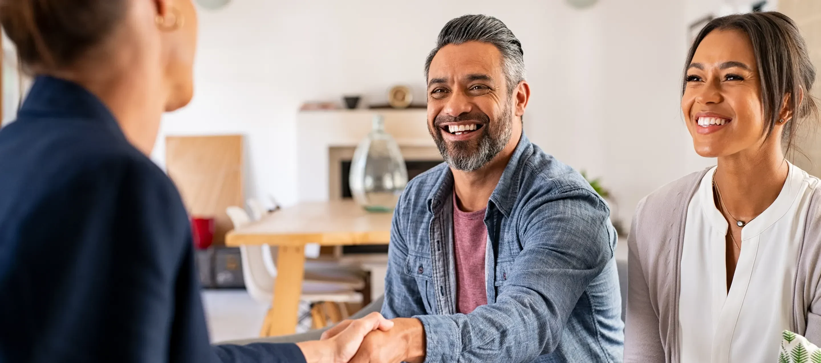 A professional meeting takes place as two people shake hands across a table in a modern, bright interior setting.